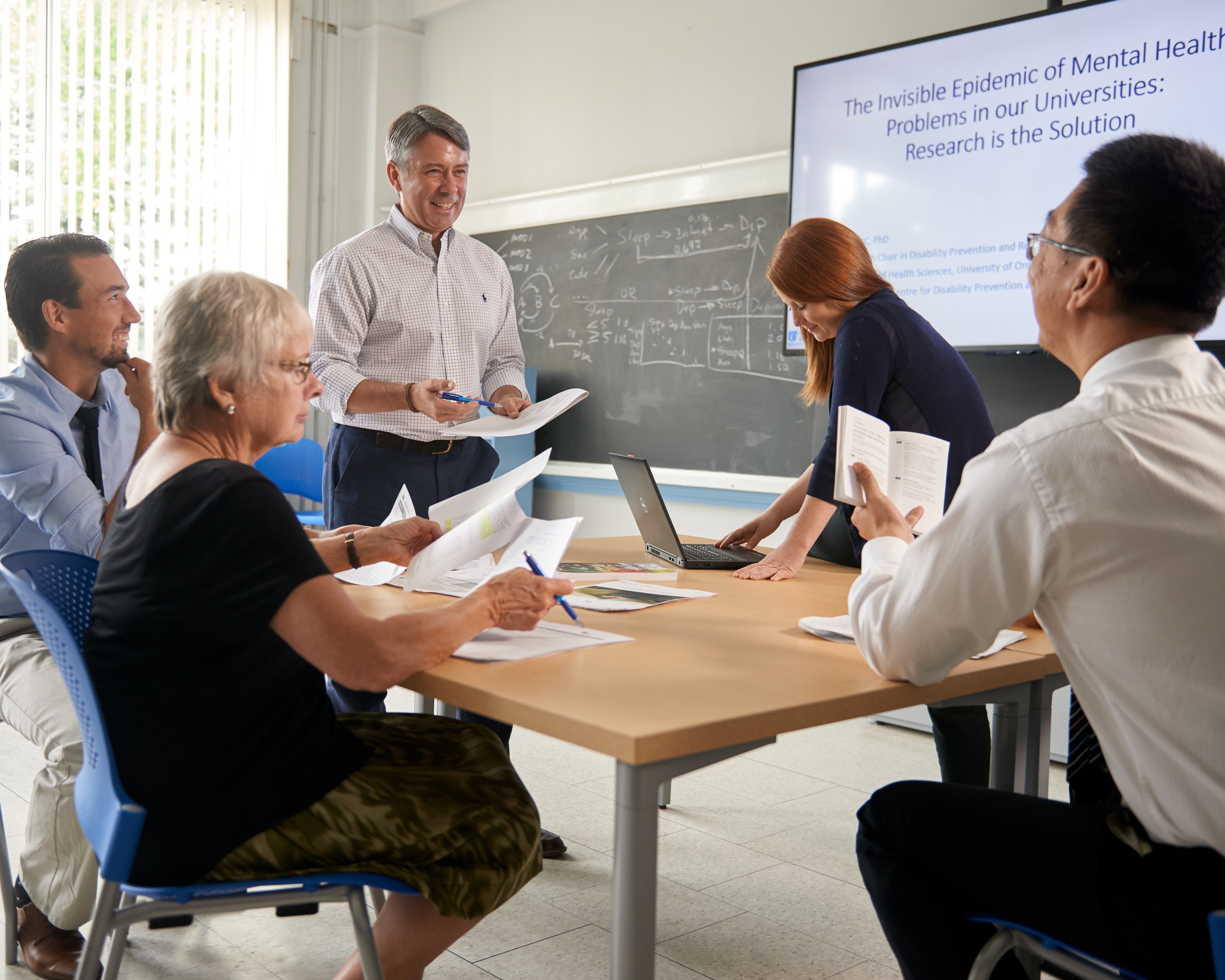   A diverse group of adults participate in a classroom workshop. Some are seated around a table with documents, while two presenters stand at the front near a screen with a mental health presentation. 